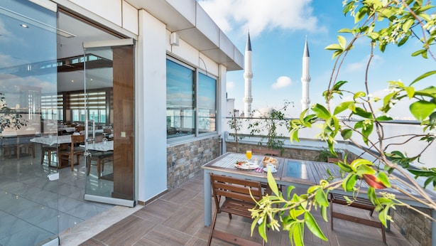 Un área de comedor al aire libre moderna con una mesa y sillas, una pared de vidrio y vistas a una mezquita con minaretes.