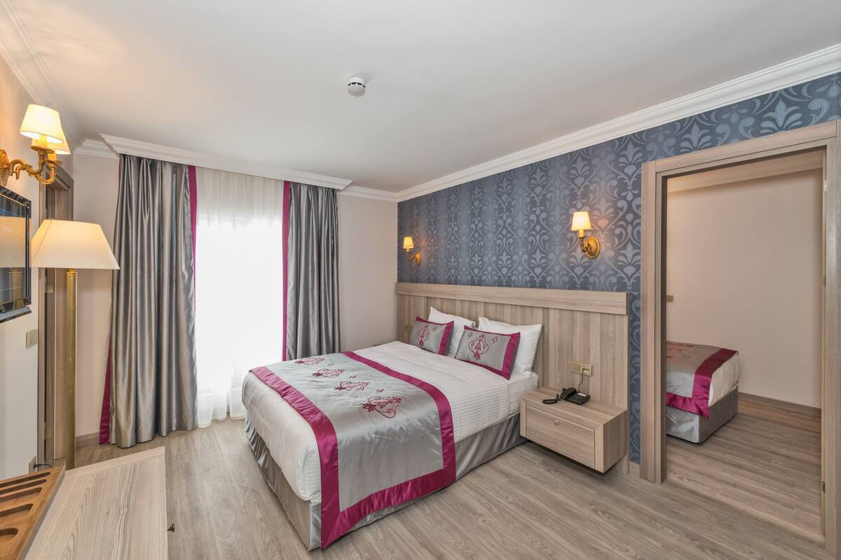 A hotel room with a large bed, bedside tables, a nightstand, a wardrobe, and a window with curtains.