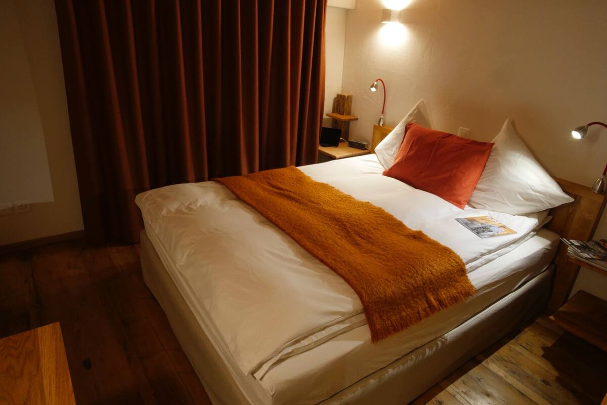 Una cama bien tendida con sábanas blancas y un plaid marrón, una mesita de noche con lámpara, y piso de madera.
