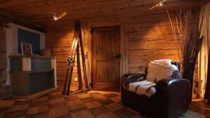 Interior de una cabaña de madera con suelo de cuadros, un sillón de cuero, una puerta de madera y una escalera apoyada en la pared.