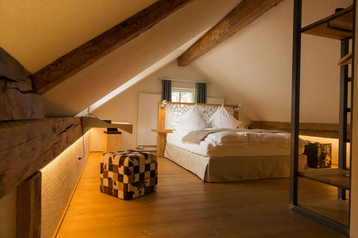 Un dormitorio acogedor en el ático, con techo inclinado, un reposapiés de cuadros, cama con ropa blanca y una ventana con cortinas.