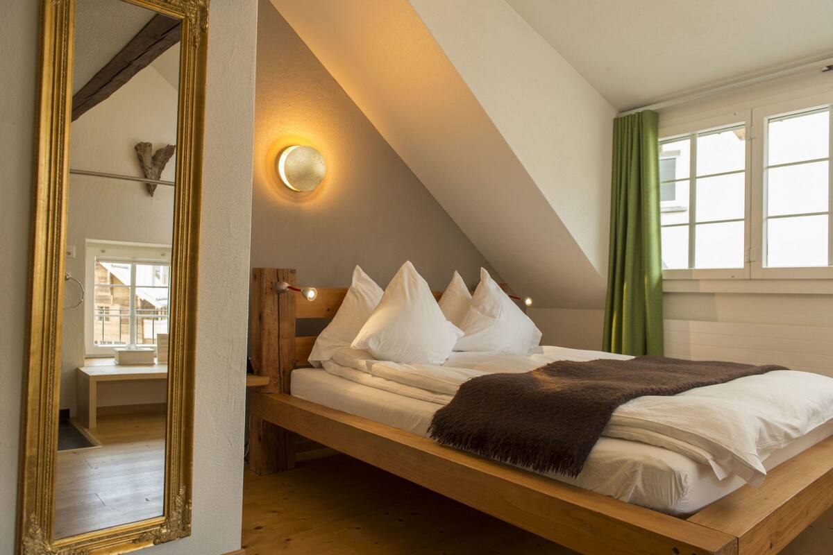 Un dormitorio con techo inclinado, una cama con ropa blanca, un cabecero de madera y una lámpara fijada a la pared.