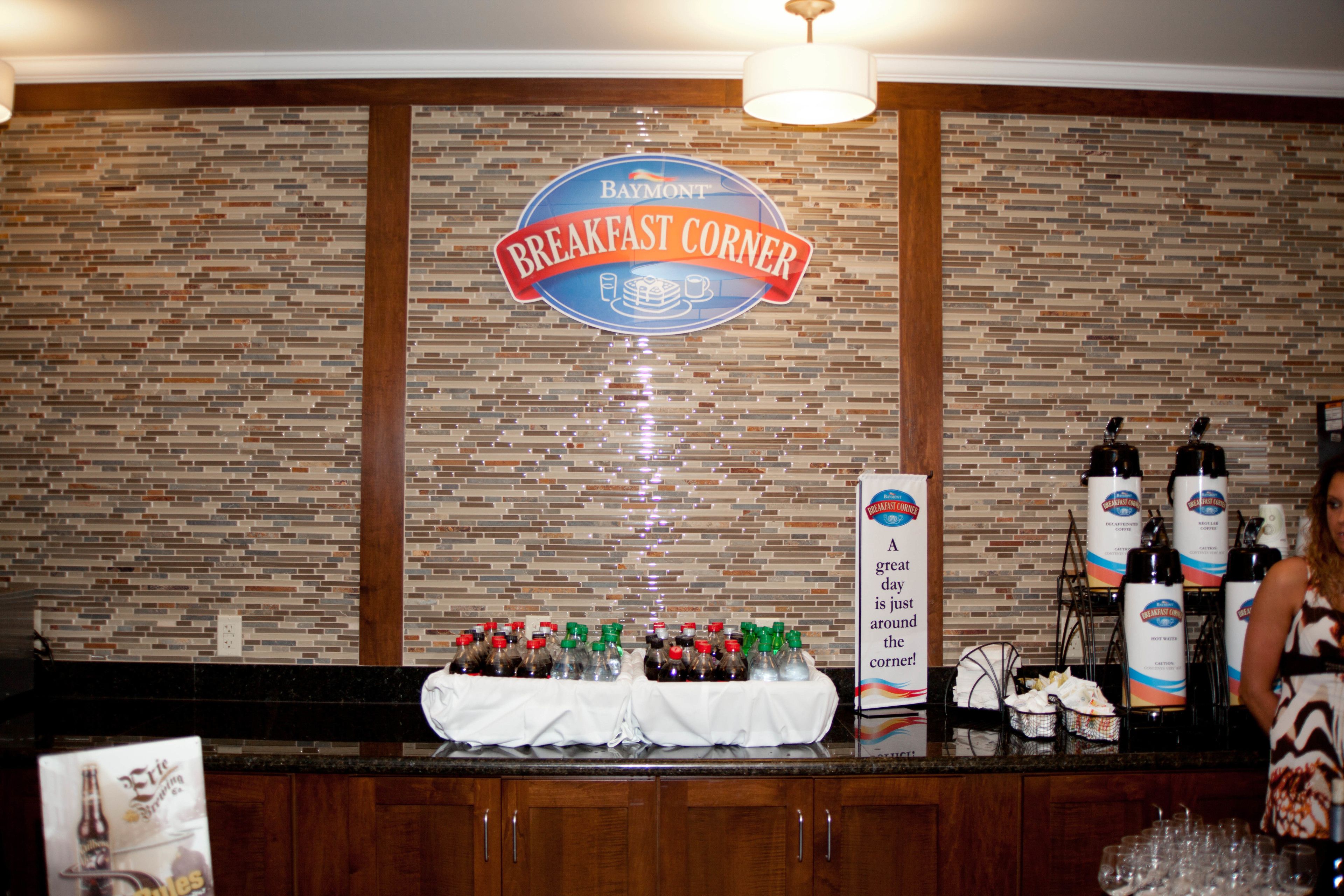 A breakfast bar area with a sign reading "Baymont Breakfast Corner," bottles of beverages, and a table with a white tablecloth.