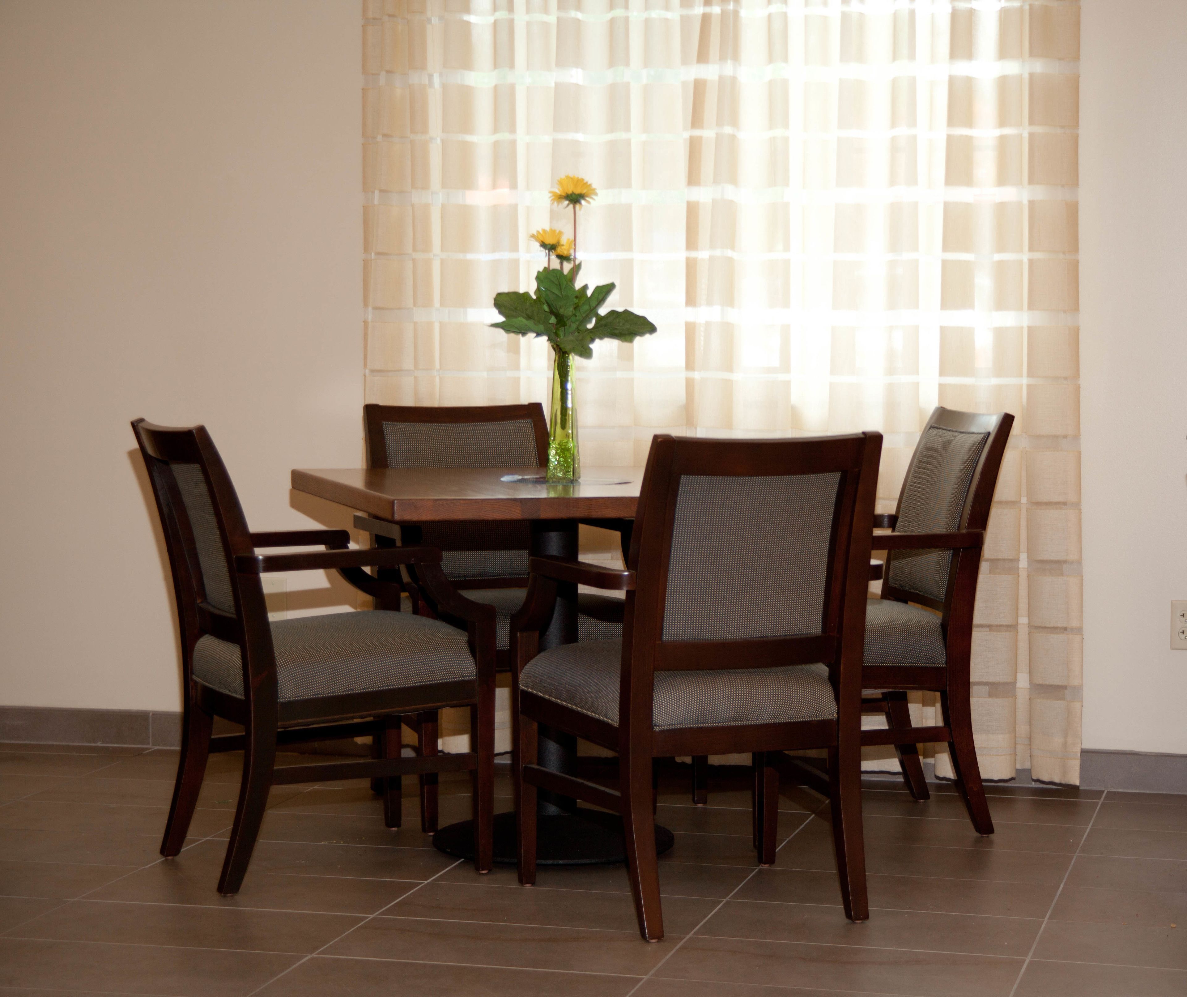 A dining table with four chairs, a vase with flowers, and a window with sheer curtains.