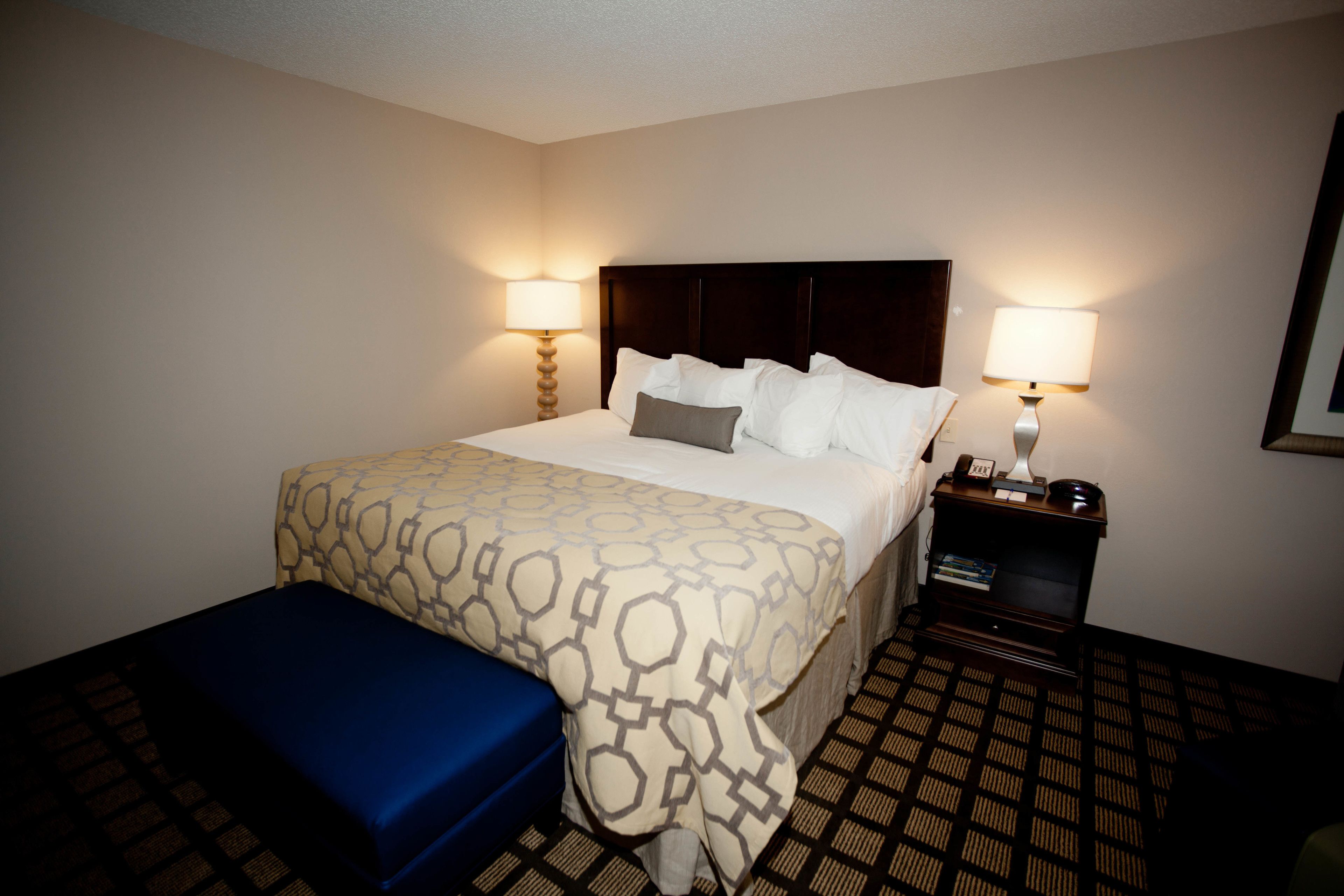 A hotel room with a large bed, two bedside lamps, a bedside table with a phone and a clock, and a blue bench.