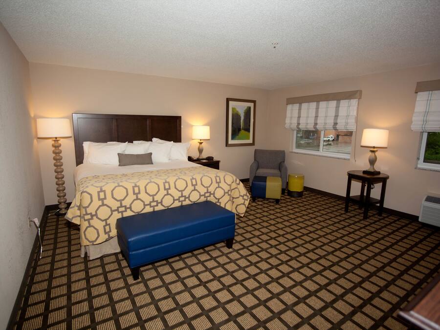 Une chambre d’hôtel comprenant un lit, deux tables de chevet avec des lampes, une chaise, une petite table et une fenêtre avec des stores.