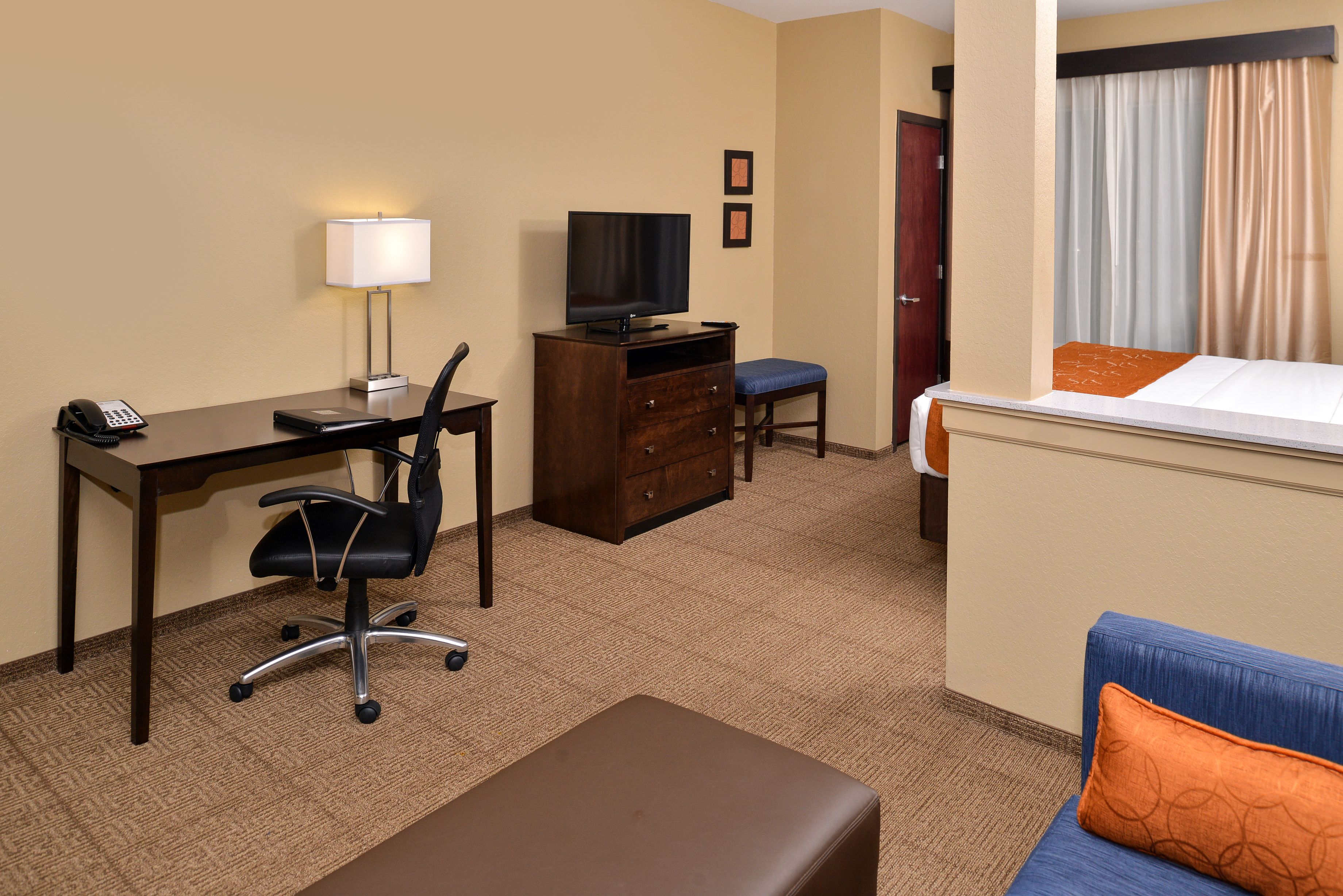 Habitación de hotel con escritorio, silla, televisión, cómoda y cama con almohada azul y naranja.