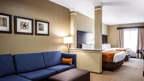 A hotel room with a blue sofa, a brown ottoman, a bed with white linens, a nightstand, and framed pictures on the wall. - Comfort Suites Northwest - Cy - Fair (Houston)
