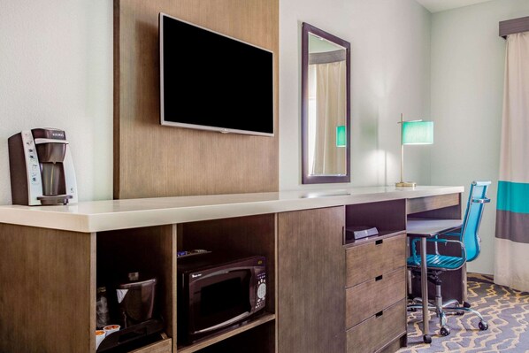 A hotel room with a flat-screen TV, a coffee maker, a microwave, a desk with a chair, and a bedside lamp.