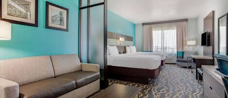 A hotel room with a sofa, a bed, a desk, and a TV. There are framed pictures on the wall.