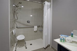 Zona de ducha con barra de apoyo, una silla blanca y repisa de vidrio con artículos de tocador.