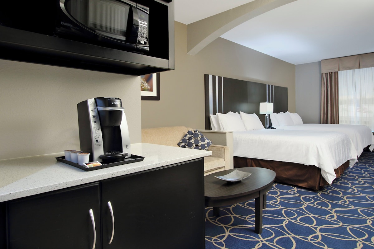 Habitación de hotel con cama, escritorio con cafetera, microondas, mesita de noche y alfombra estampada.
