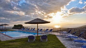 A poolside area with lounge chairs and umbrellas, overlooking a body of water and mountains during sunset.