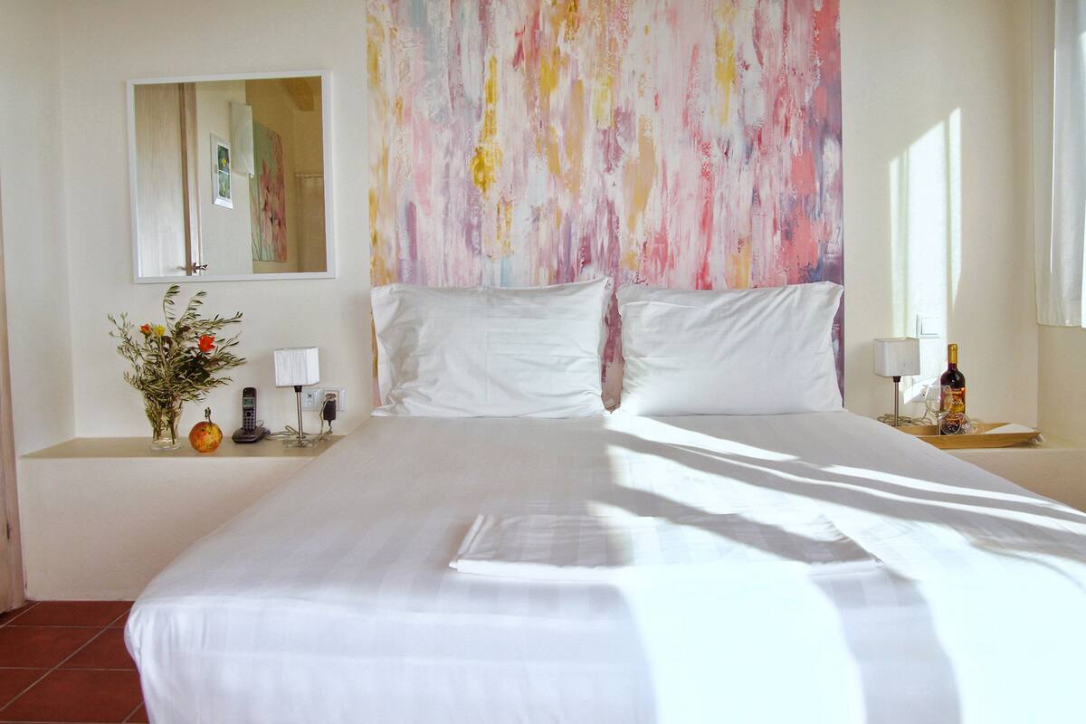 A hotel room with a large bed, a bedside table with a lamp, a vase with flowers, and a bottle of wine on the nightstand.