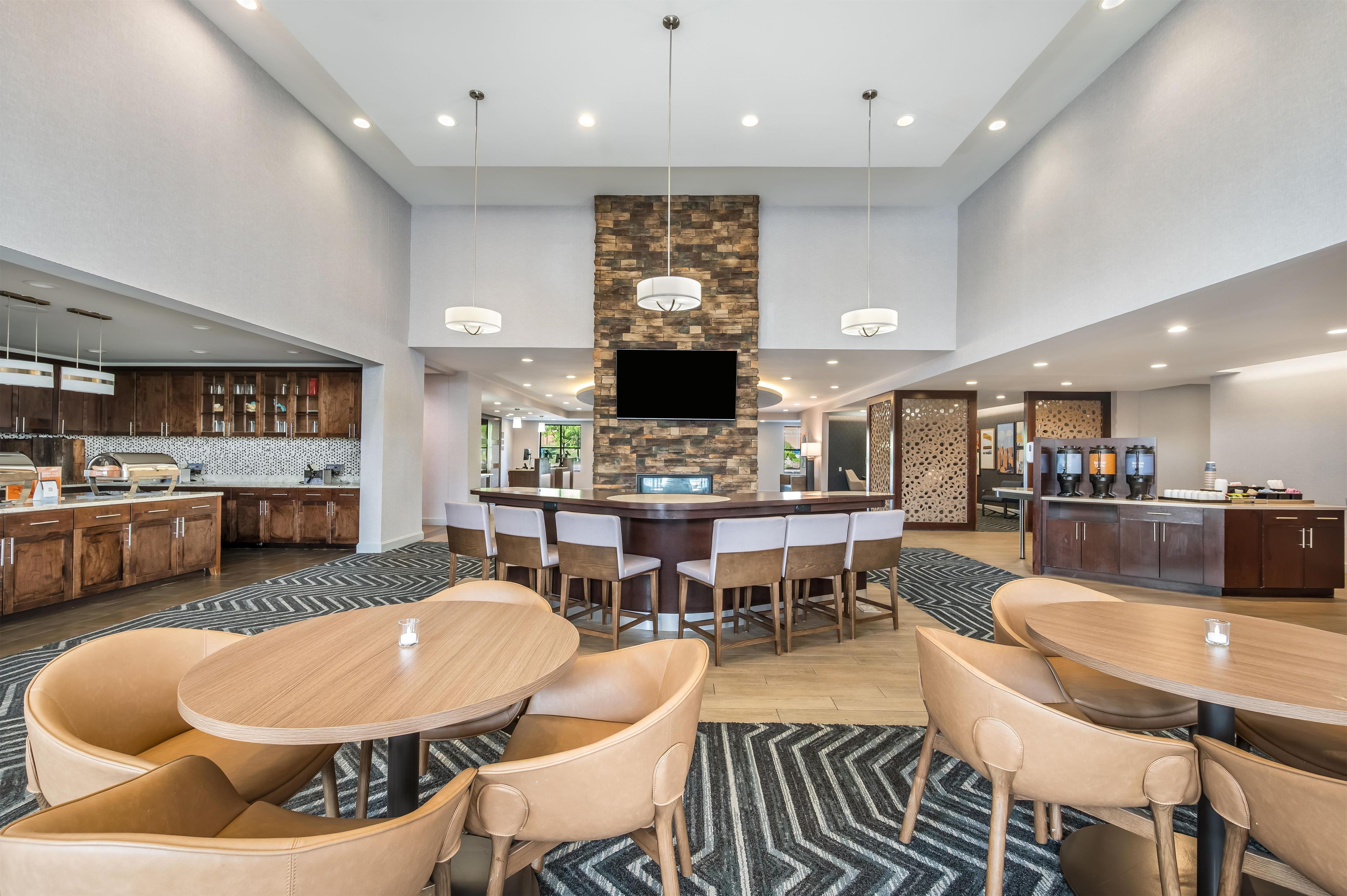 A modern hotel lobby with a central bar, seating area, and a kitchen in the background.