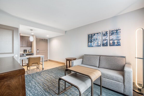 A modern living room with a grey sofa, a wooden coffee table, and a patterned rug. There is a kitchen area with cabinets and a dining table. - Homewood Suites By Hilton Cincinnati Mason (Mason)