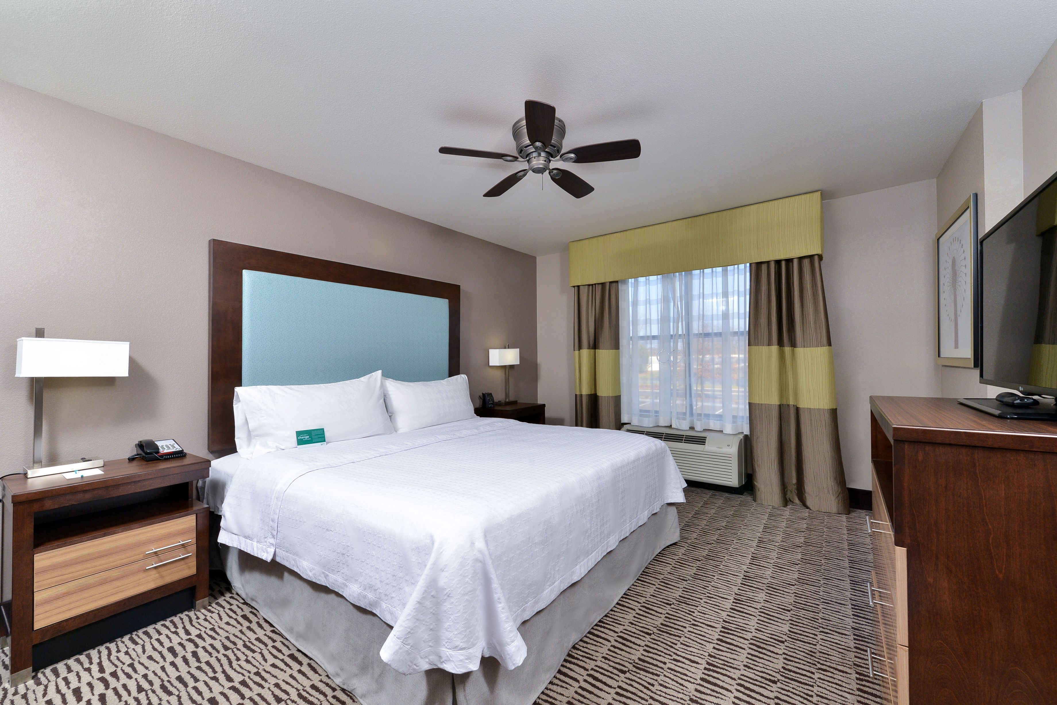A hotel room with a bed, bedside tables, a desk, a television, and a ceiling fan.
