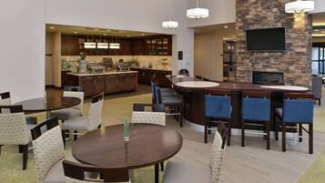 Un lobby de hotel moderno con una zona de bar central, mesas de comedor y una pared decorativa de piedra con un televisor de pantalla plana.