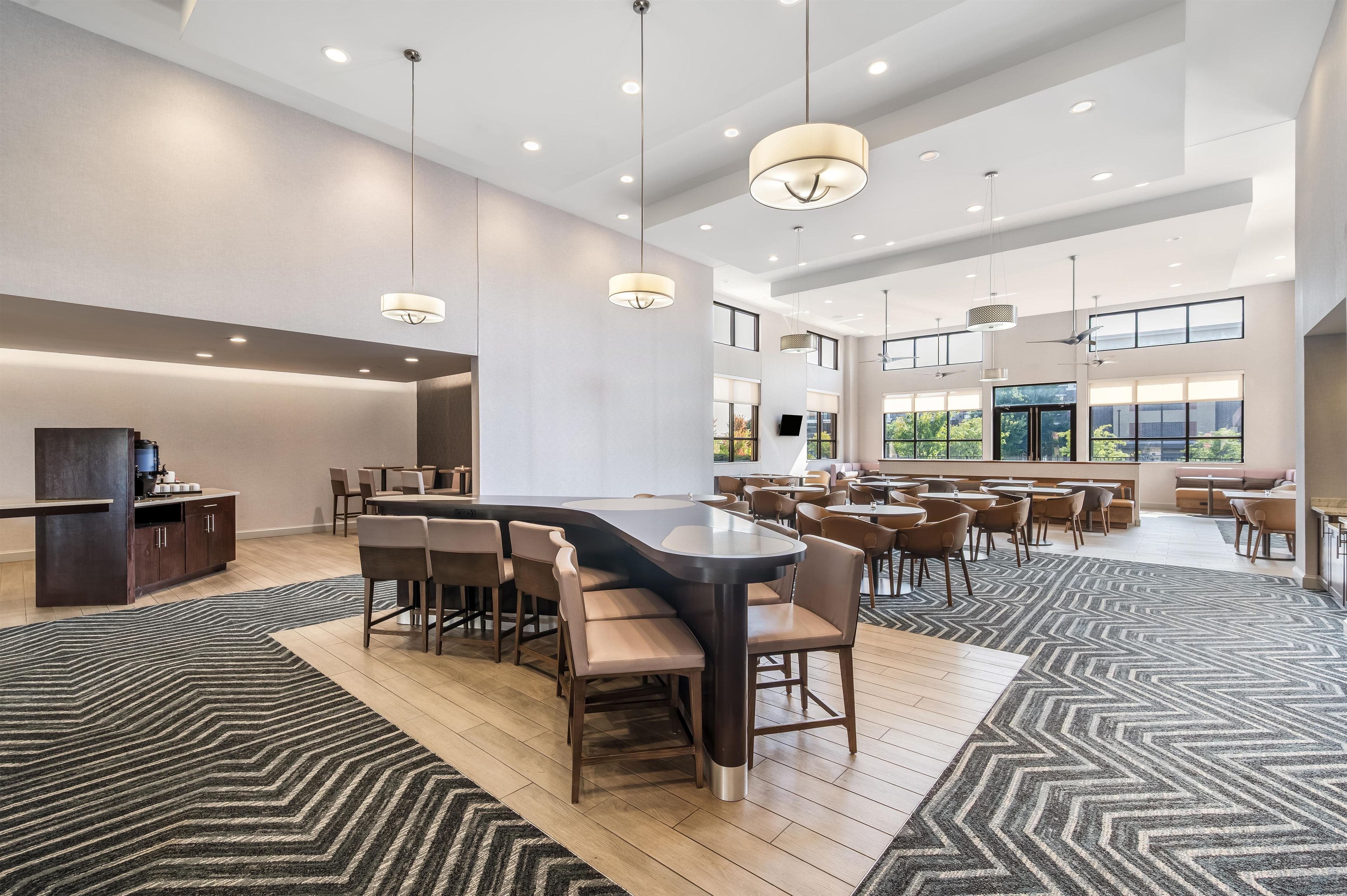 A modern hotel lobby with a central bar area, multiple seating options, and large windows providing natural light.