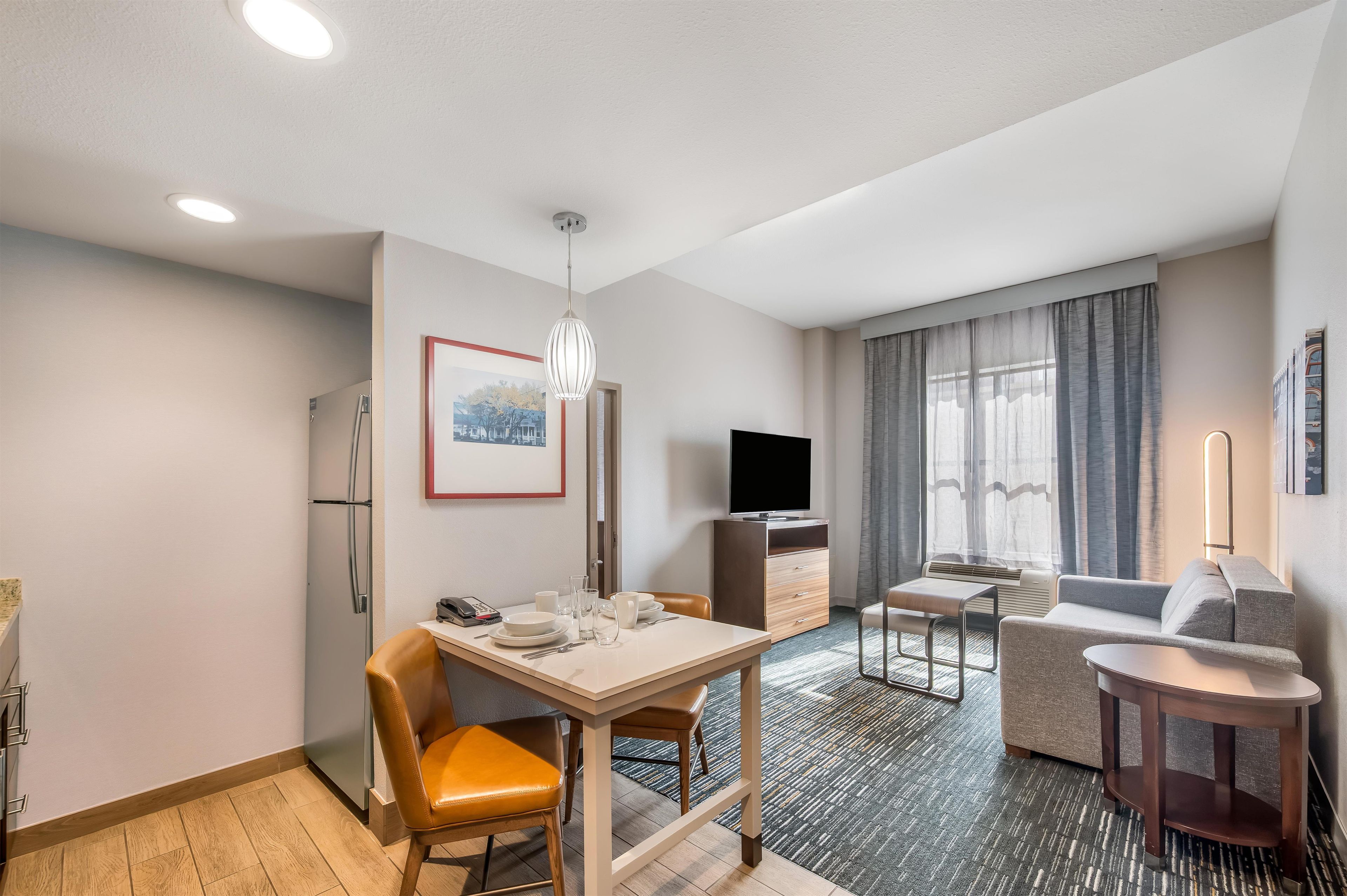 Et moderne hotelværelse med spiseplads, en sofa, et fjernsyn og et lille bord med lampe.