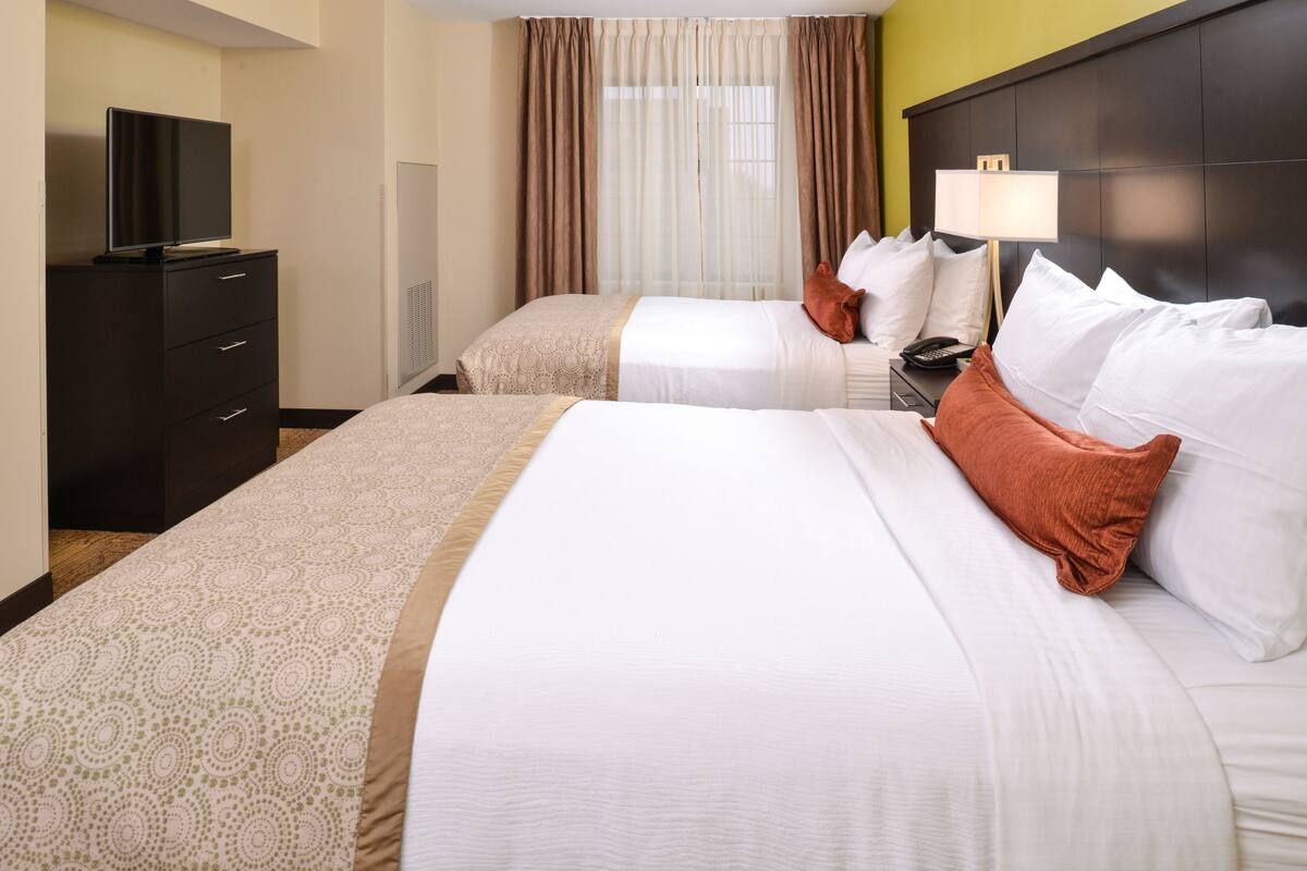 A hotel room with two beds, a TV on a dresser, a lamp, and a window with curtains.