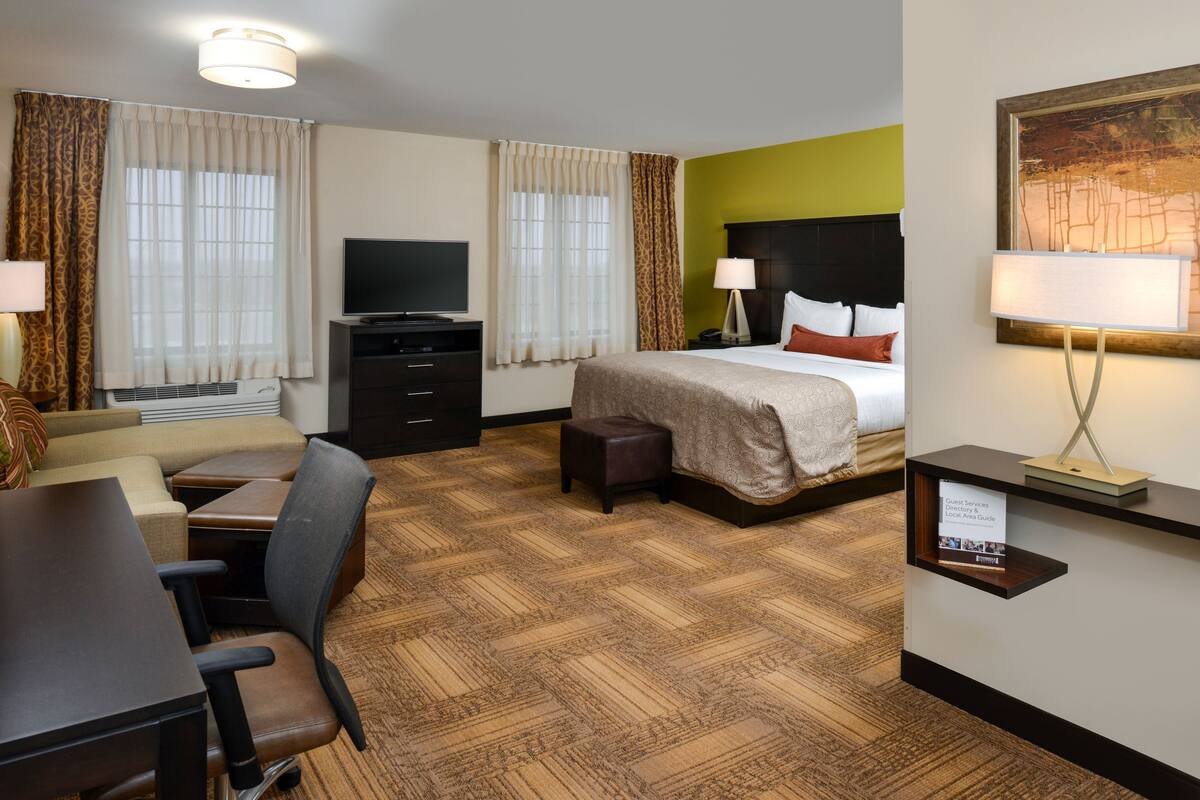 A hotel room with a bed, a desk with a chair, a television, a small table, a sofa, and a wall-mounted shelf with a sign.