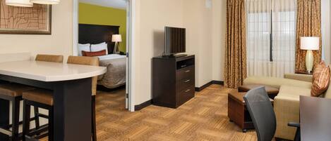 A hotel room with a dining area, a bed with a green accent wall, a TV, a desk, and a sofa.