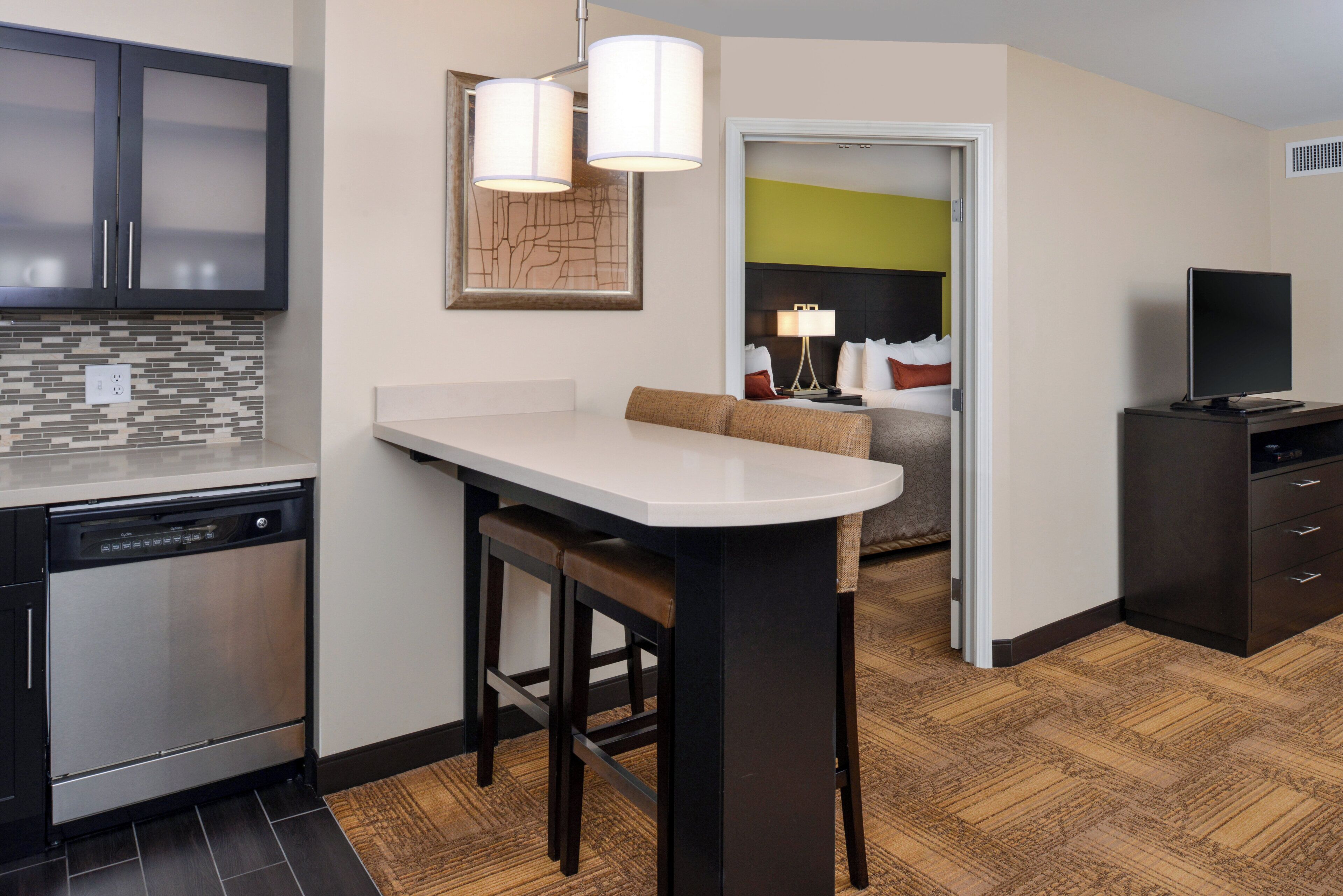 A modern kitchen with a built-in dishwasher, a dining area with a bar stool, and a living space with a sofa and a television.