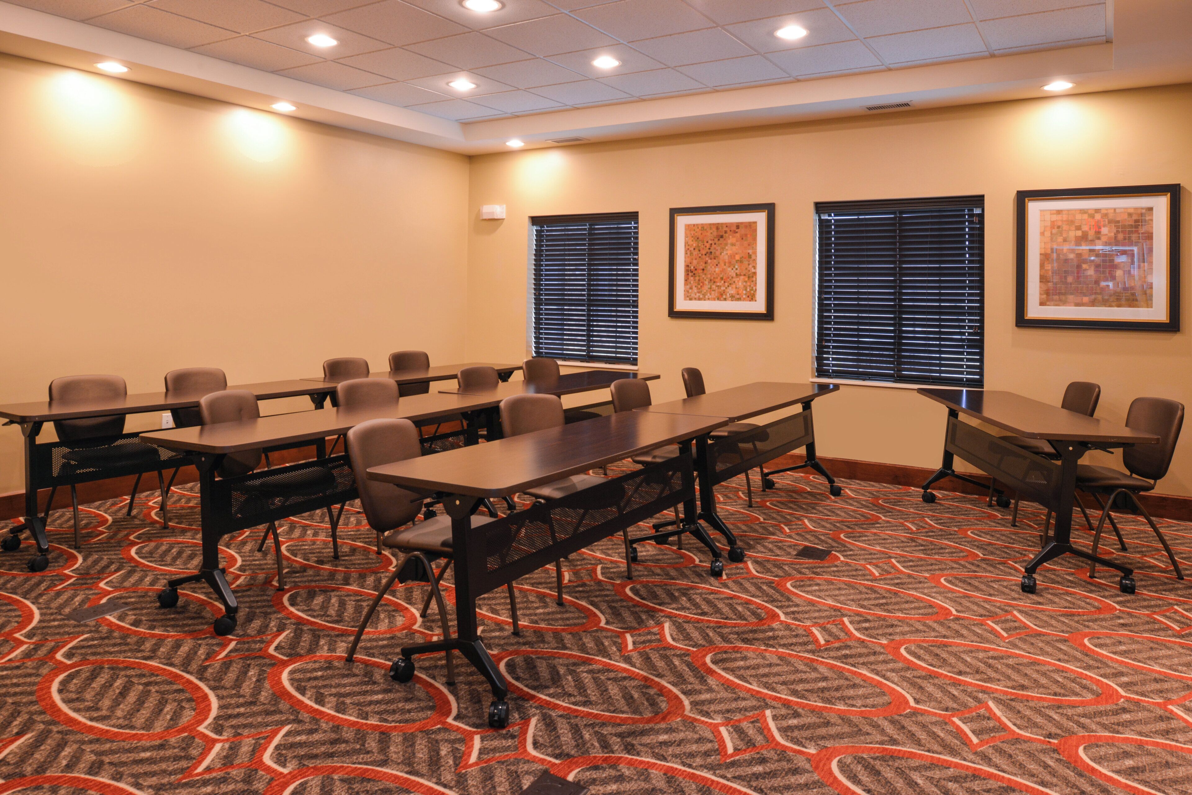 A conference room with multiple tables and chairs, carpeted floor, ceiling lights, and framed artwork on the walls.