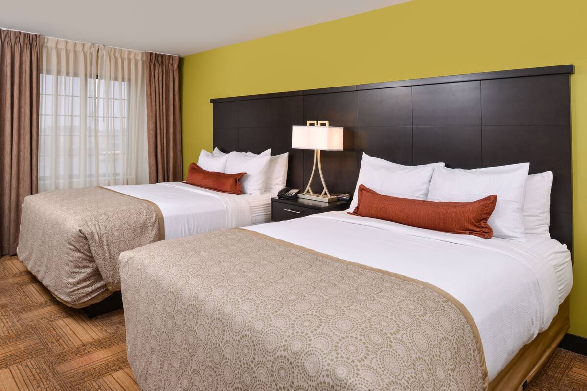 A hotel room with two beds, a nightstand, a lamp, and a window with curtains.