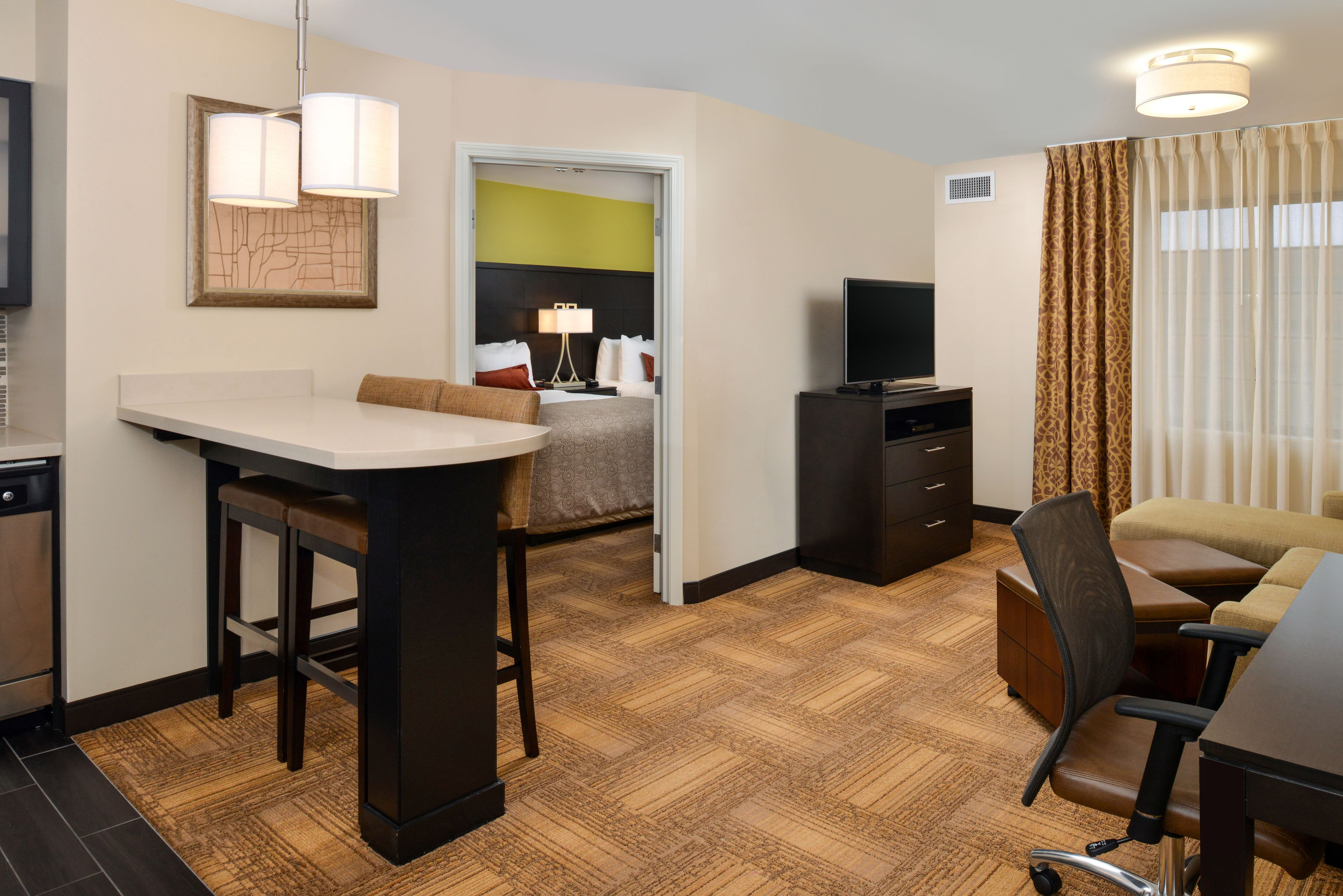A hotel room with a kitchenette, a dining area with a bar stool, a seating area with a sofa and armchairs, a television, and a large window with curtains.