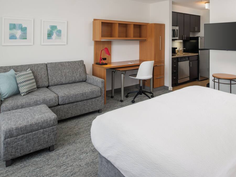Moderni hotellihuone, jossa on harmaa sohva, valkoinen sänky, työpöytä tuolilla, pieni keittiöalue ja litteänäyttöinen televisio.