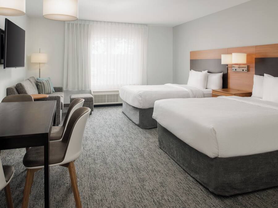 Quarto de hotel com uma cama grande, uma cama menor, escrivaninha com cadeiras, televisão de tela plana e janela com cortinas.