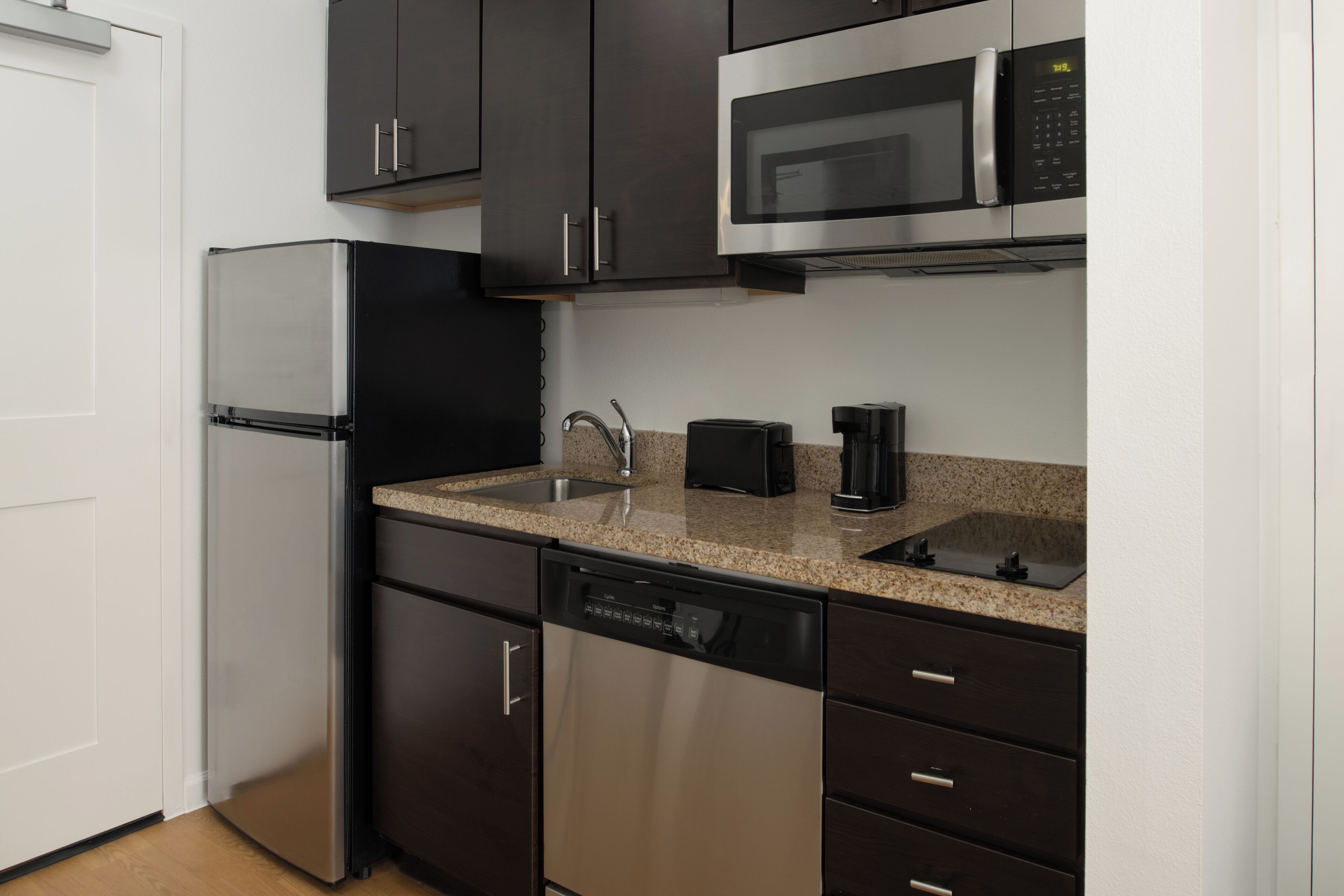 A compact kitchen with dark cabinets, a stainless steel refrigerator, a microwave, a dishwasher, and a granite countertop.