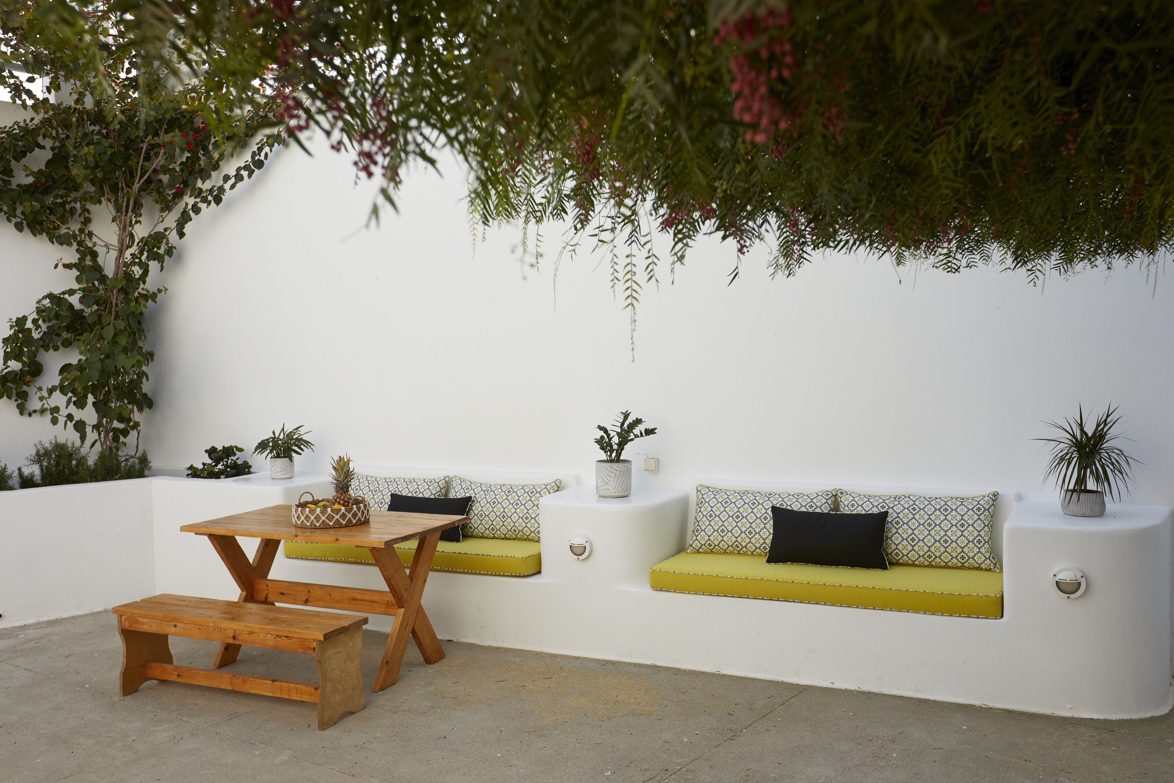 A patio with a wooden table and benches, a white wall, and potted plants.