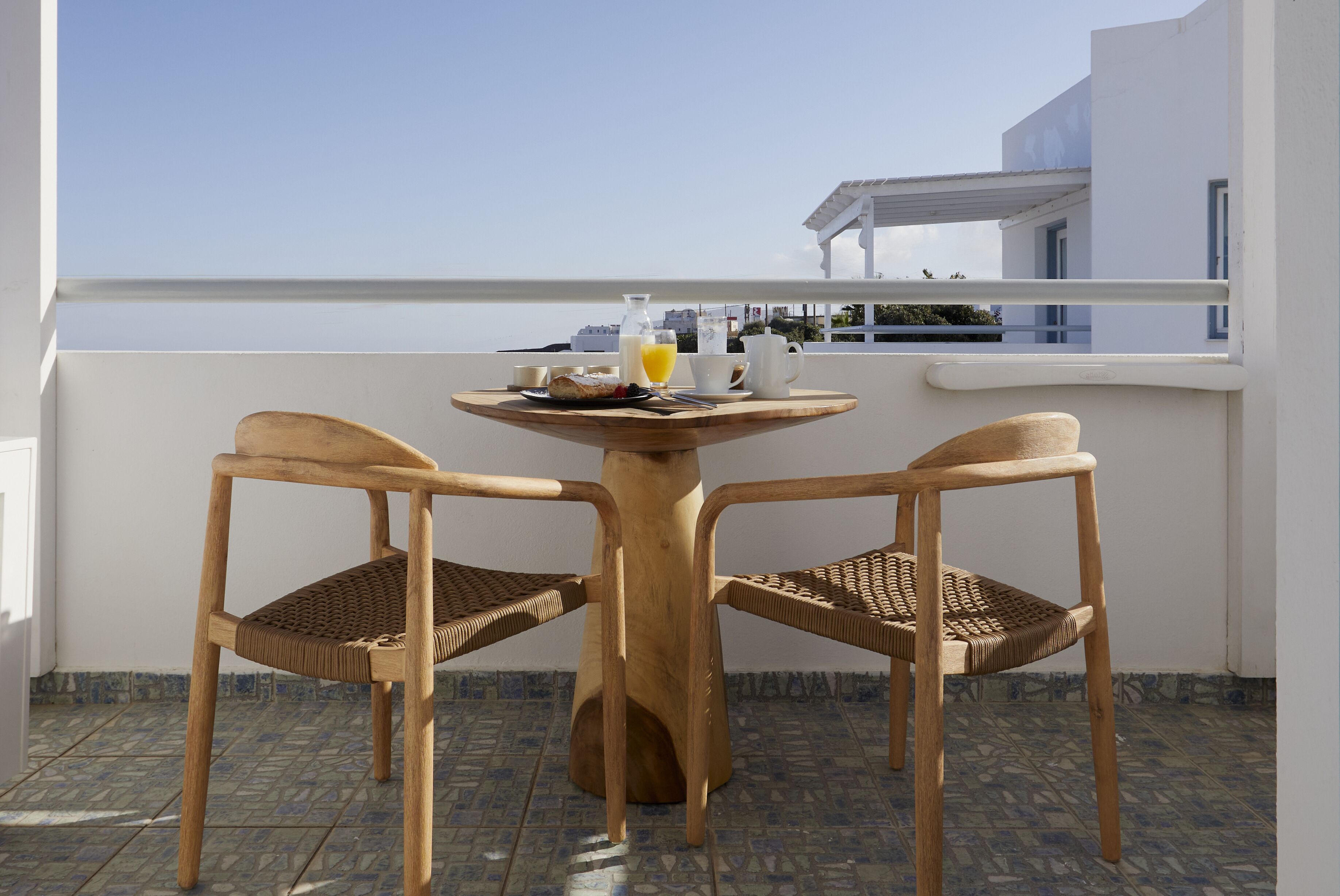 Une table en bois, accompagnée de deux chaises, sur un balcon doté d’un sol carrelé et d’une rambarde blanche.