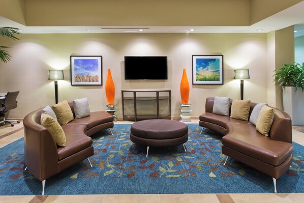 A hotel lobby with a curved brown sofa set, a round ottoman, a flat-screen TV, two orange vases, and a patterned carpet.