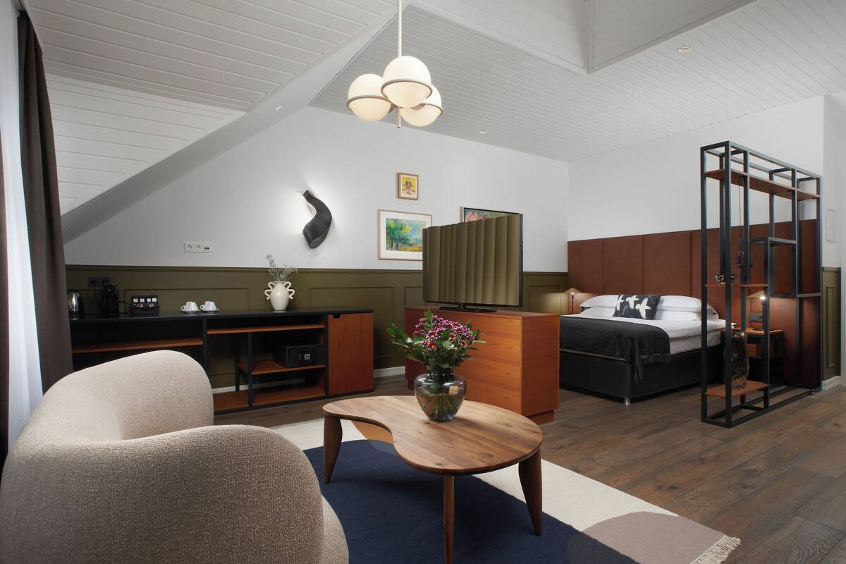 A modern hotel room with a lofted ceiling, a bed with a wooden frame, a desk with a chair, a coffee machine, and a vase with flowers.
