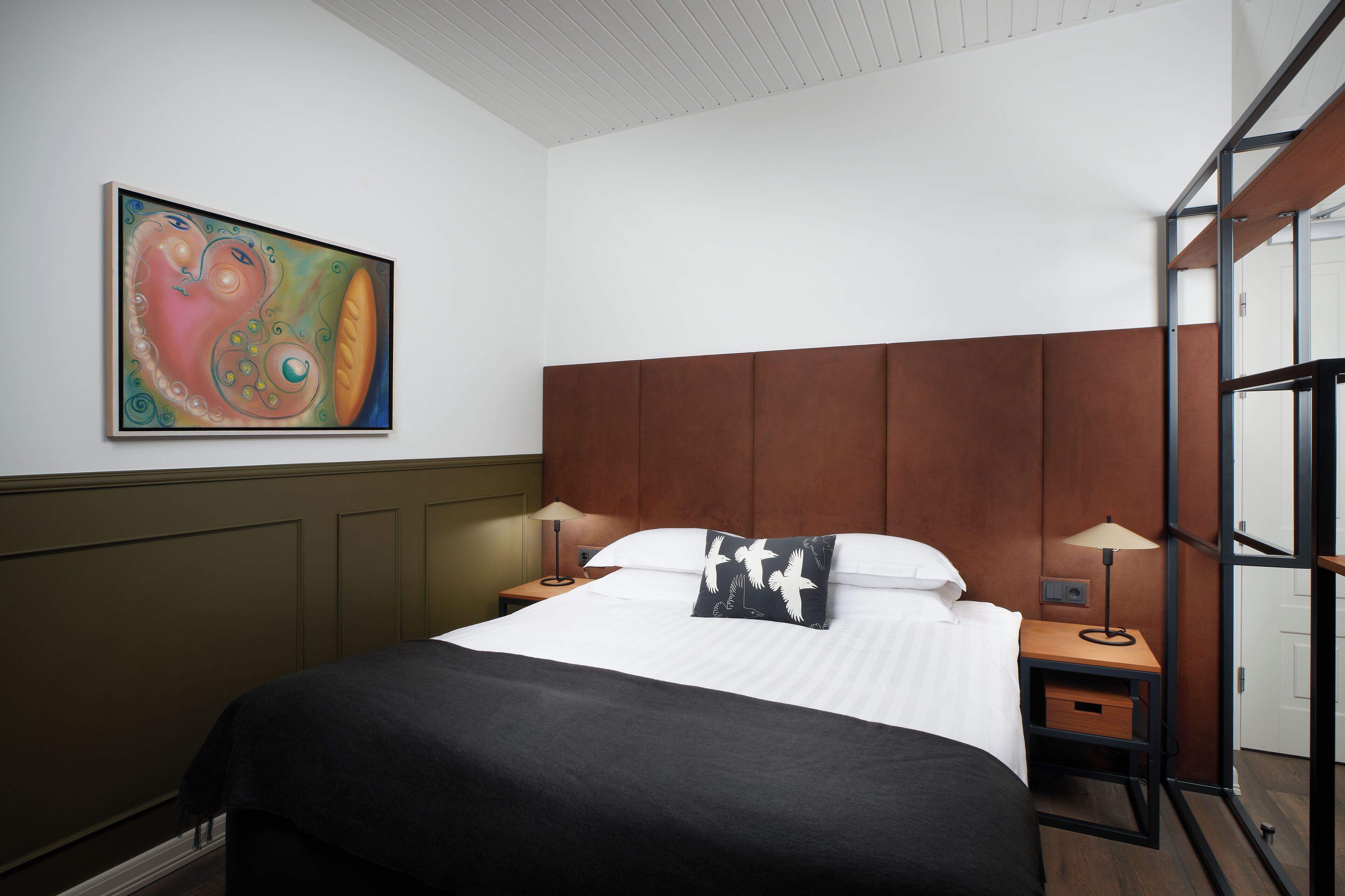 A neatly made bed with a white and black bedspread, two bedside tables with lamps, a wooden headboard, and a colorful abstract painting on the wall.