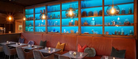 Un restaurant avec des banquettes et des tables, des étagères illuminées de lumière bleue où sont disposés des bocaux et des bouteilles, et des lampes suspendues.