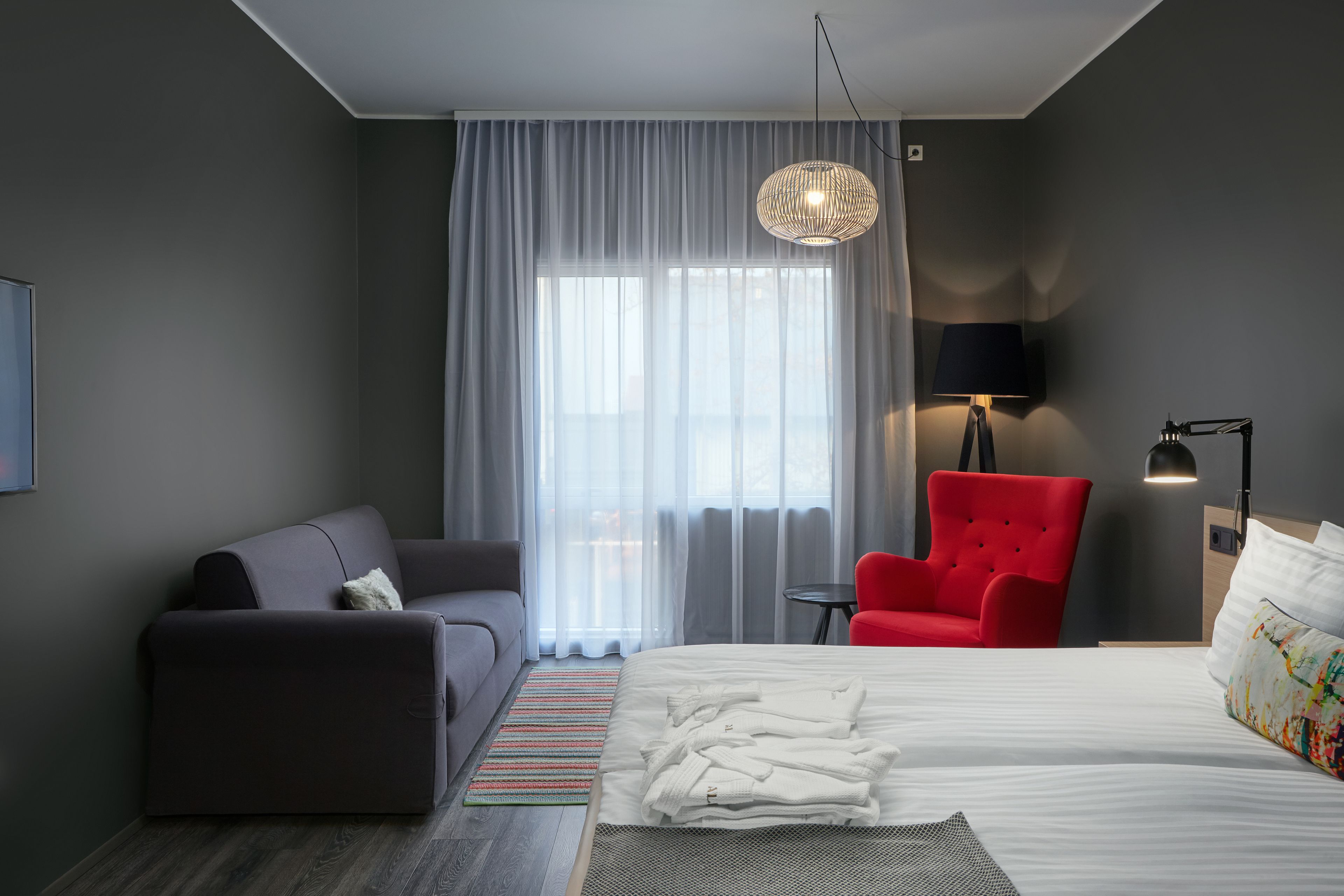 Una habitación de hotel moderna con una cama, un sillón rojo, un sofá gris y un ventanal con cortinas translúcidas.