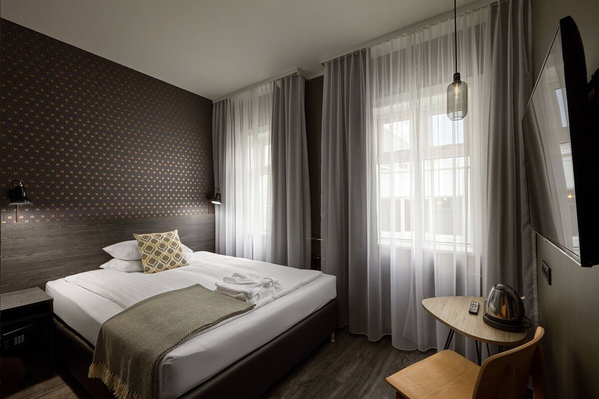 A hotel room with a neatly made bed, a small table with a kettle, and a TV on the wall.