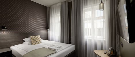 A hotel room with a neatly made bed, a small table with a kettle, and a TV on the wall.