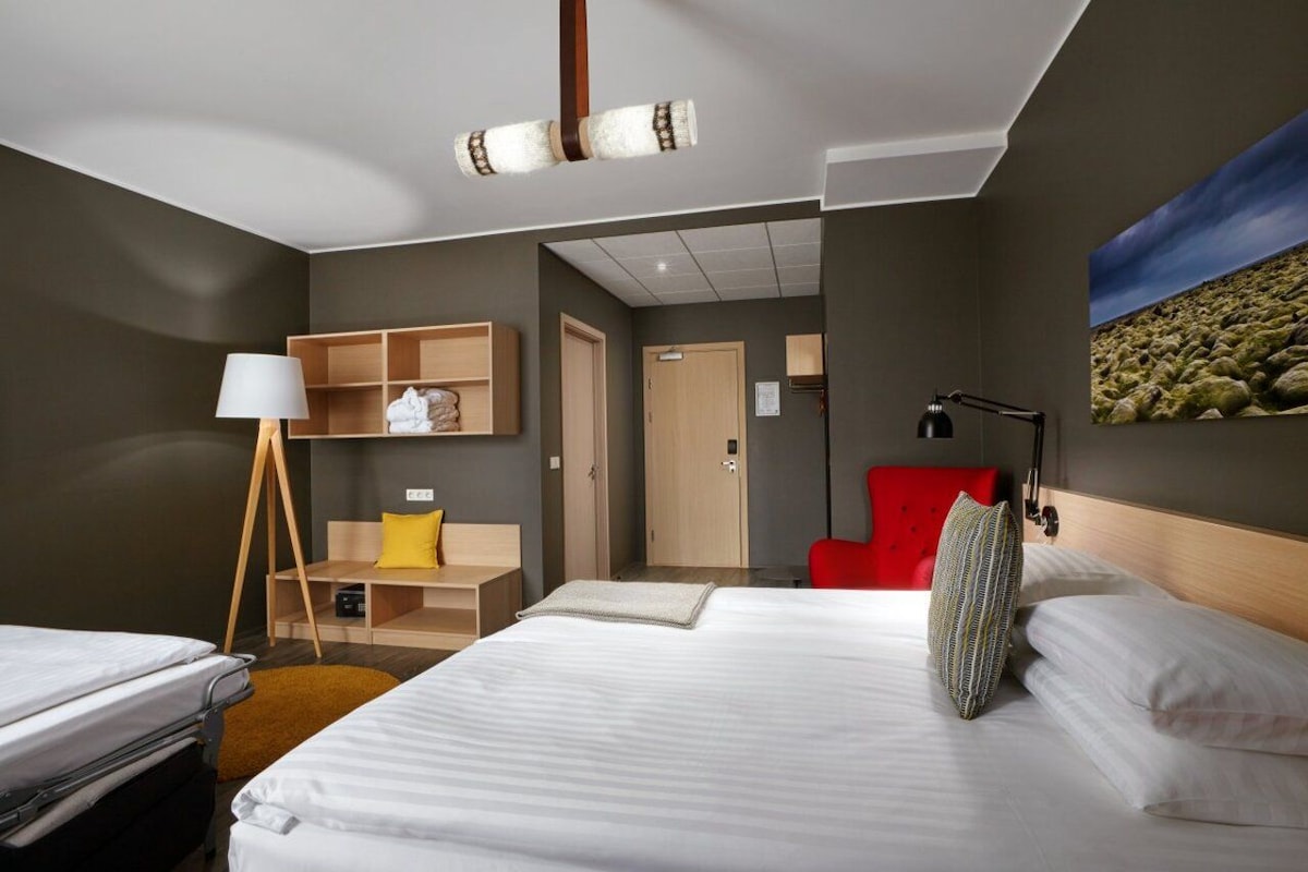 Habitación de hotel con cama, mesita de noche, lámpara y una silla roja.