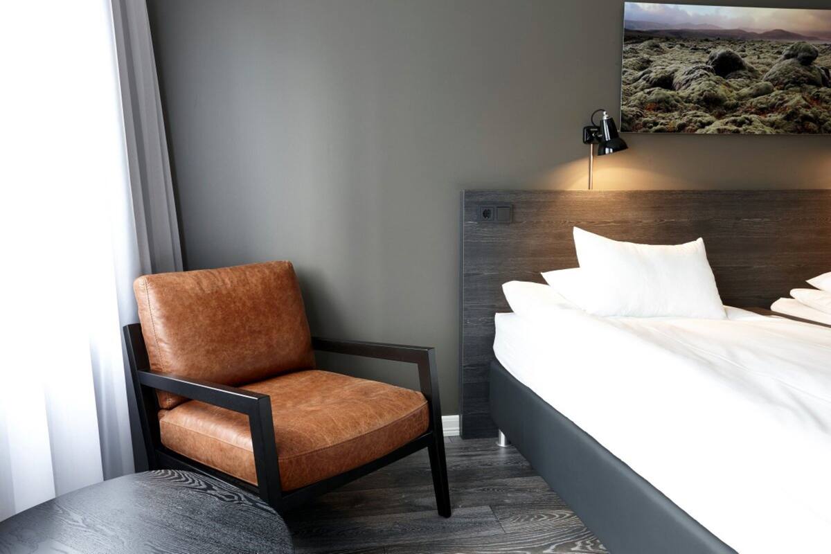 A modern hotel room with a single bed, a brown leather chair, a round table, and a wall-mounted lamp.