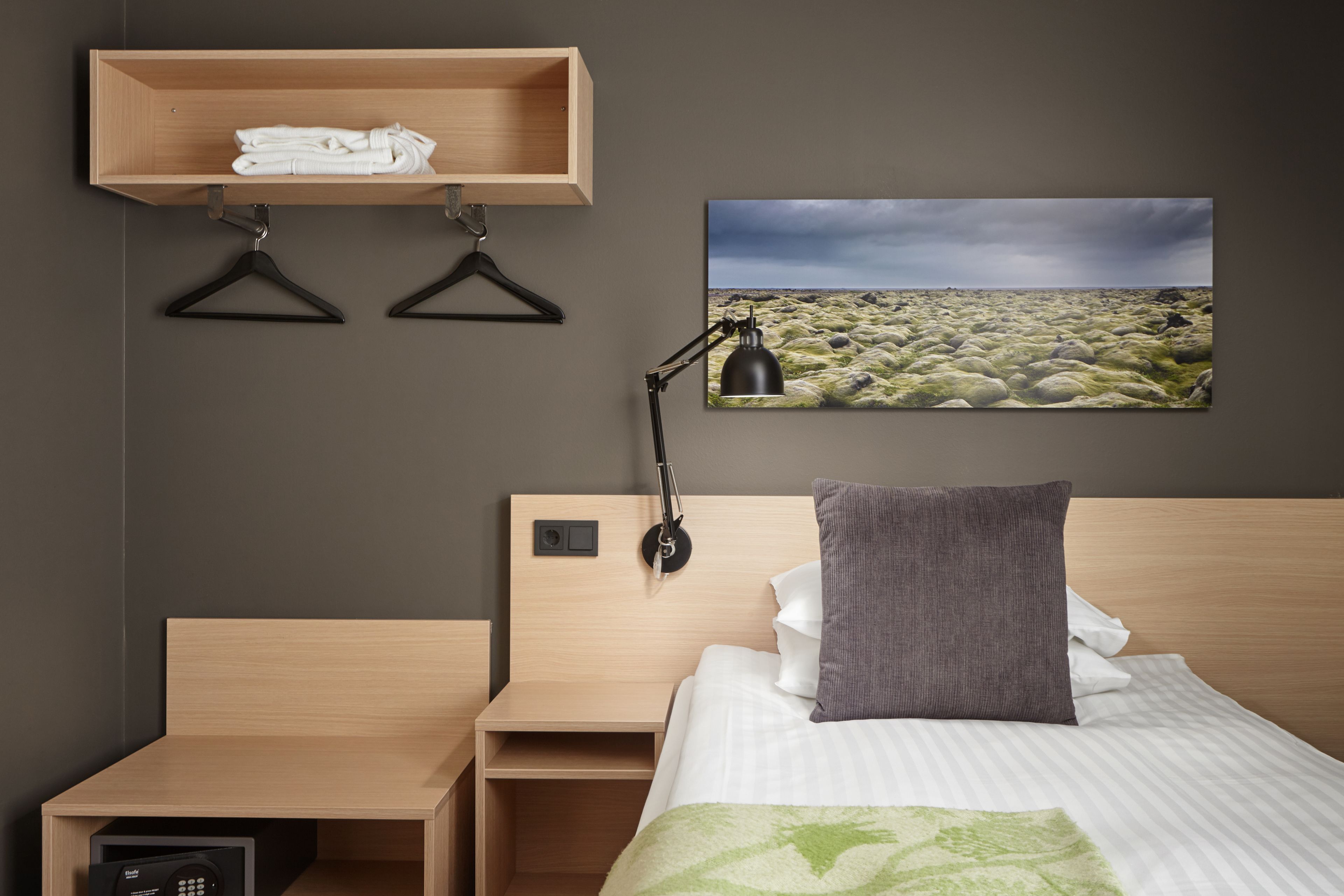 A hotel room with a bed, a bedside table, a nightstand, a wooden shelf with towels, a black lamp, and a landscape painting on the wall.
