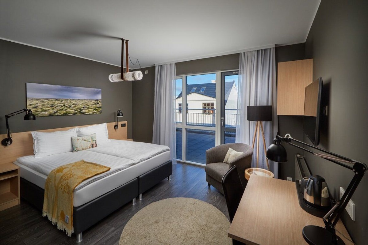 Habitación de hotel con cama, escritorio, silla, televisión y balcón con cortinas.