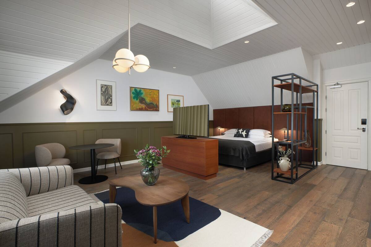 A modern hotel room with a loft-style ceiling, a bed, a seating area with a sofa and chairs, a small table with a vase, and artwork on the walls.