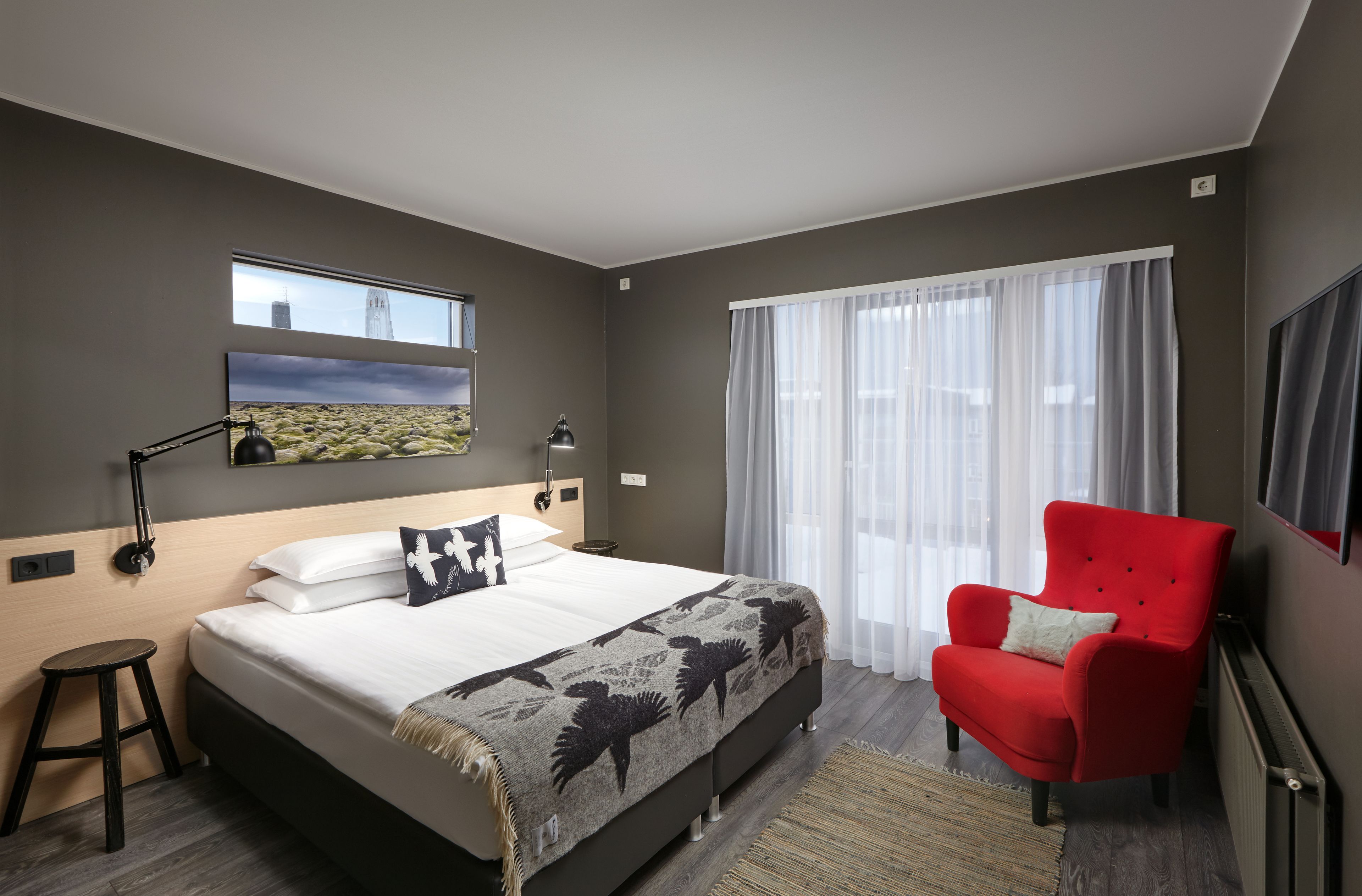 Quarto de hotel moderno com uma cama grande, uma poltrona vermelha, uma janela com vista para a cidade e uma televisão na parede.