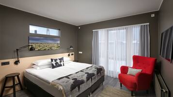 Quarto de hotel moderno com uma cama grande, uma poltrona vermelha, uma janela com vista para a cidade e uma televisão na parede.